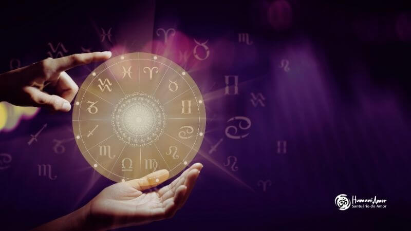 O despertar da alma Close-up de duas mãos interagindo com um mapa astral circular e translúcido que brilha com uma luz dourada suave. Uma mão sustenta o mapa por baixo, enquanto um dedo da outra mão aponta para um dos setores. Ao fundo, diversos símbolos astrológicos flutuam em um ambiente desfocado com iluminação roxa e pontos de luz (bokeh). No canto inferior direito, aparece o logotipo branco "HumaniAmor - Santuário do Amor"